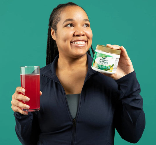 A smiling woman in a navy zip-up jacket holds a glass of red juice in one hand and Weight Manager drink mix powder in the other, set against a solid teal background.