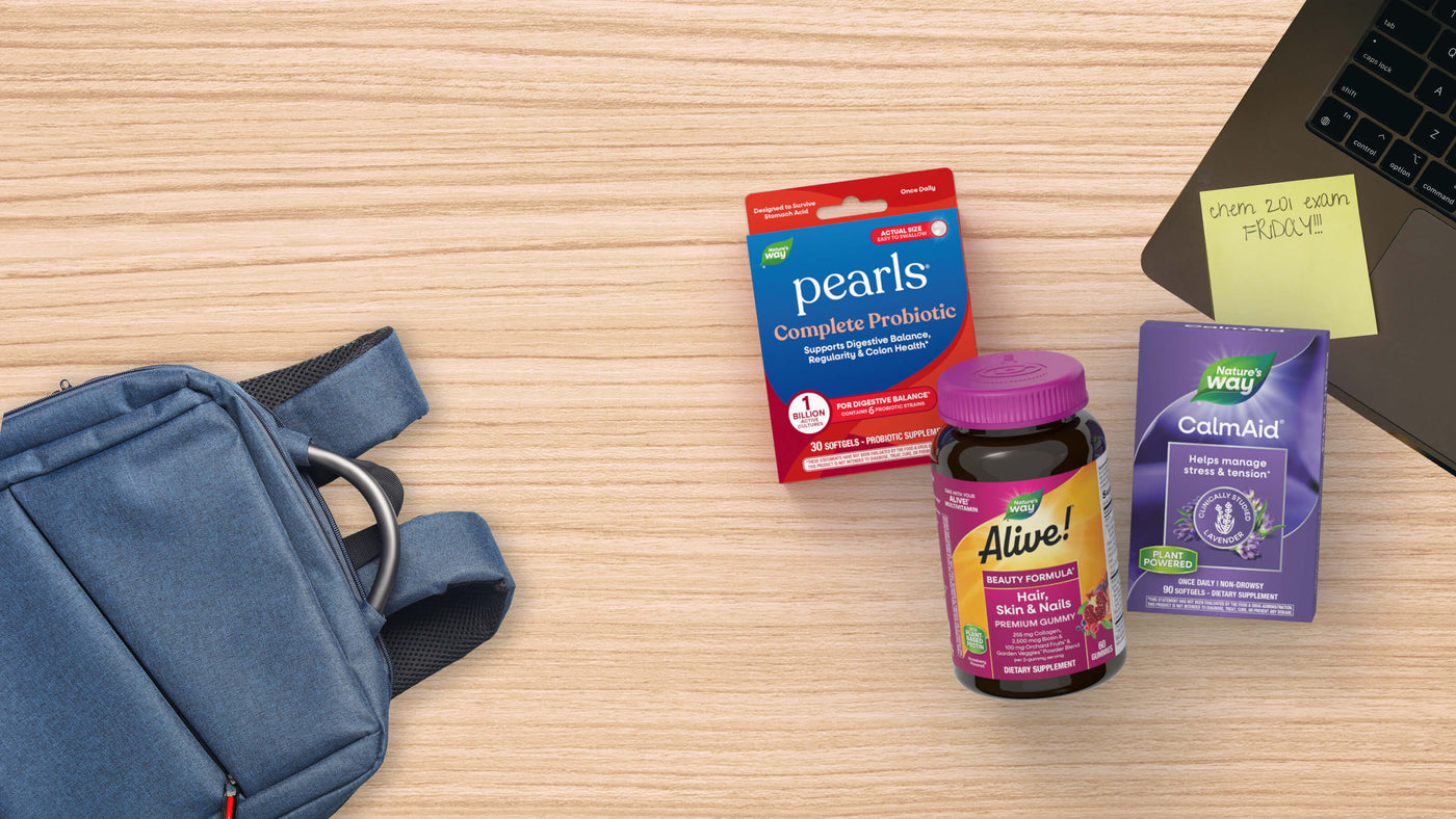 A blue shoulder bag lies on a light wood desk alongside three supplement products: a red and blue Pearls Complete Probiotics box, a purple bottle of Alive Hair Skin and Nails, and a purple Way CalmAid box. A laptop corner and sticky note are also visible.