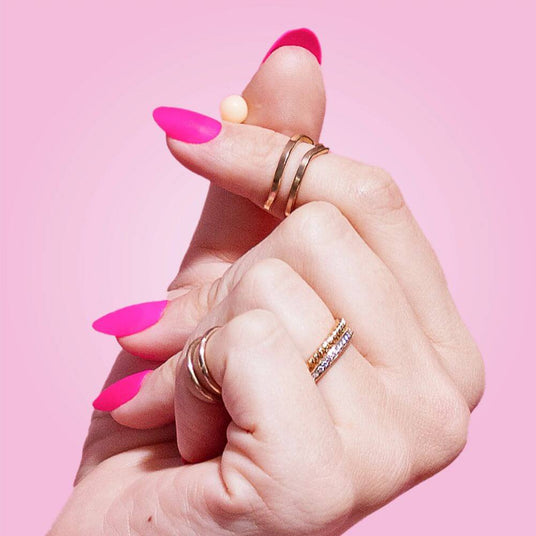 Close-up of a hand holding a probiotic Pearl softgel with pink nail polish and multiple gold rings on a pink background