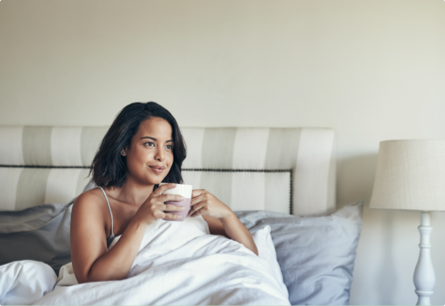 <{%DETAIL2_15794%}>A woman sitting up in bed under a white blanket with a mug in her hands.
