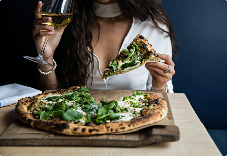 A woman with a glass of white wine in one hand and a slice of spinach pizza in the other hand, the rest of the pizza is in the foreground.