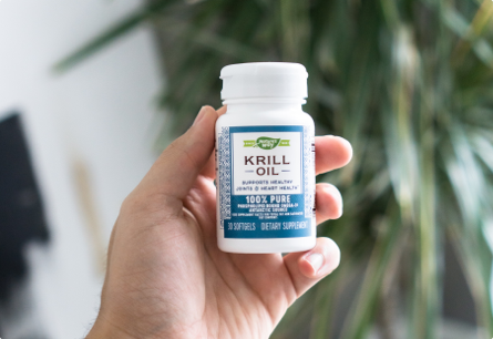 A hand holding a bottle of Krill Oil with a green, leafy plant in the background.