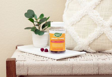<{%DETAIL1_14262%}>A bottle of Nature's Way Immune Power gummies on a tray with two gummies to the left of the bottle with a small plant and a throw pillow in the background.