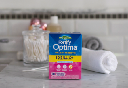 A package of Fortify Optima Women's Probiotic on a bathroom counter next to a jar of cotton swabs and a towel.
