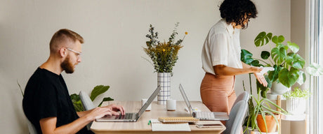 Sideview of a man working on a laptop on a table across from another open laptop while a woman tends to a plant in the corner.
