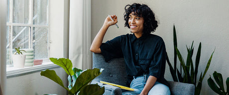 A woman surrounded by green plants sitting by a window with a pad of paper in her lap.
