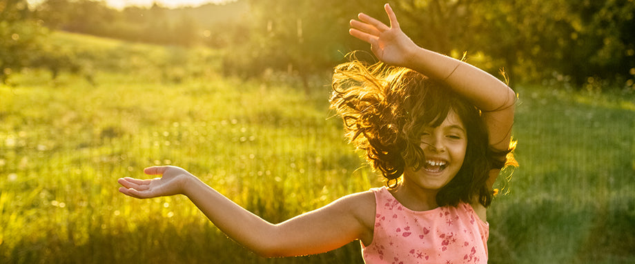 A young girl smiling in a green field with her arms in the air and her hair blowing in the wind.