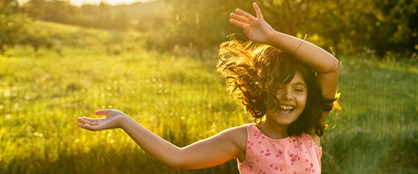 A young girl smiling in a green field with her arms in the air and her hair blowing in the wind.