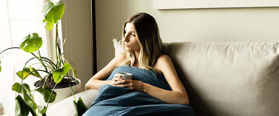 A woman sitting under a blanket on a couch next to a plant and a window.