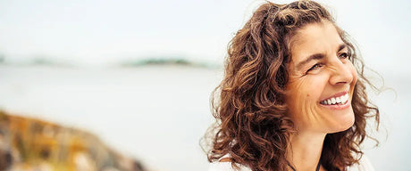 A woman smiling outdoors with a body of water and trees in the background.
