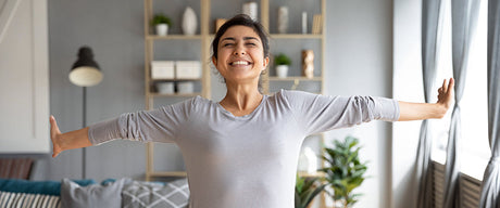 A woman smiling and stretching in her living room with the sun shining through the windows.