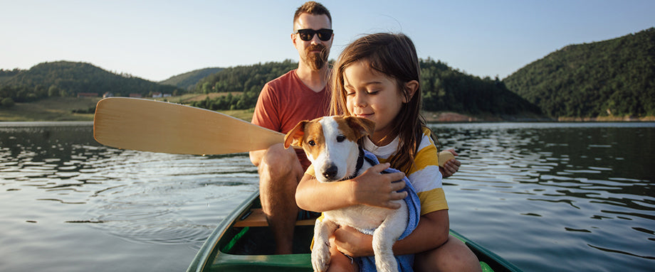 A man and a child holding a puppy sitting in a canoe on a body of water with hills in the background.