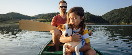 A man and a child holding a puppy sitting in a canoe on a body of water with hills in the background.