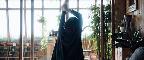 A rear view of a woman sitting in bed and stretching her arms against the sunlight.