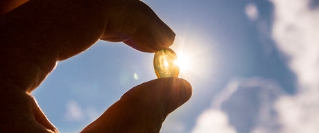 A hand holding a vitamin D softgel towards the sky with the sun shining through.