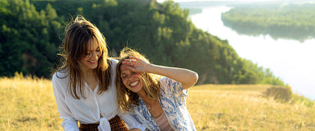 Two women laughing outside in a field with trees and a river in the background.