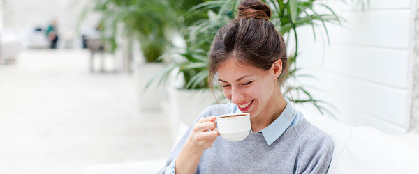 A woman smiling into a mug in front of a white wall with a leafy green plant behind her.