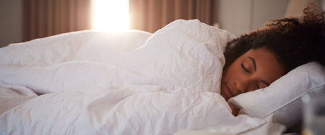 A woman sleeping under white blankets with the sun peeking through closed curtains behind her.