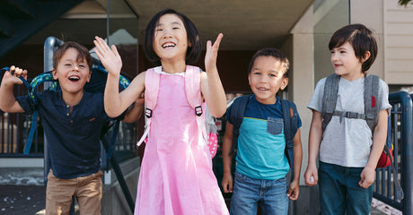 Four kids with backpacks smiling and laughing outside with their school in the background.