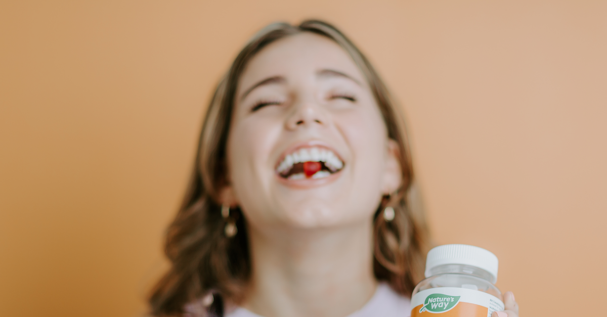 A smiling woman holding a bottle of Nature's Way Mood Lift gummies with a gummy between her teeth.