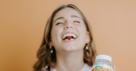 A smiling woman holding a bottle of Nature's Way Mood Lift gummies with a gummy between her teeth.