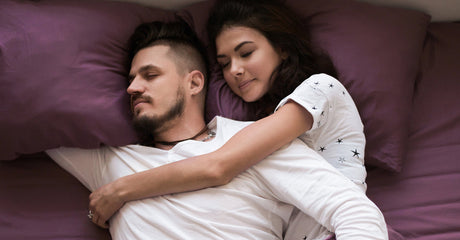 A man and a woman sleeping next to each other on purple sheets and pillows.