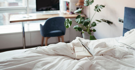 A book sitting on a bed with a pair of glasses, there's a desk in the background with a computer and a leafy plant.