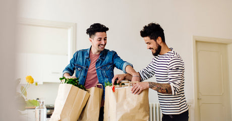 Two men smiling and opening paper grocery bags together in a kitchen.