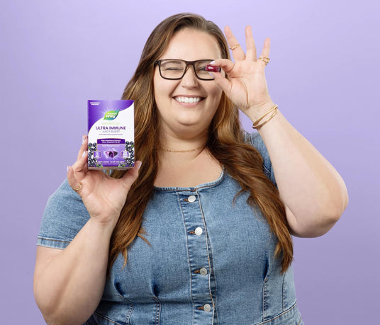 Woman holding a Nature's Way Sambucus Ultra Immune Juicy Burst supplement box against a purple background