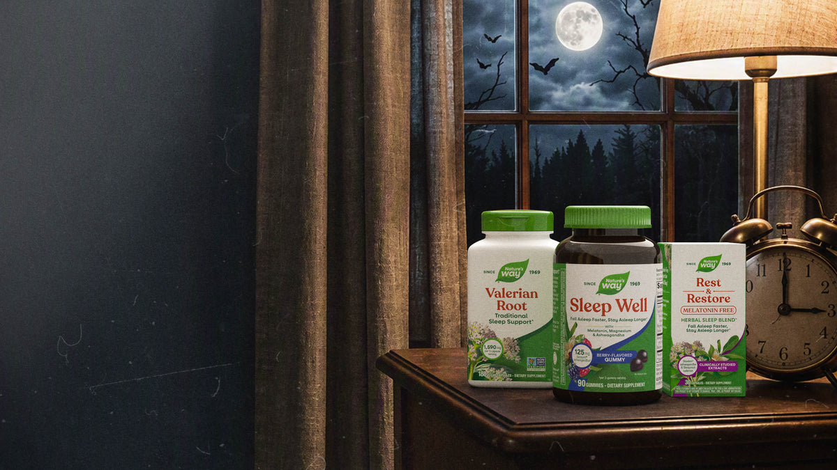 Three sleep supplement bottles are displayed on a wooden table in a cozy, atmospheric room setting. The background features a window showing a full moon and bats, a lit table lamp, and a vintage clock.