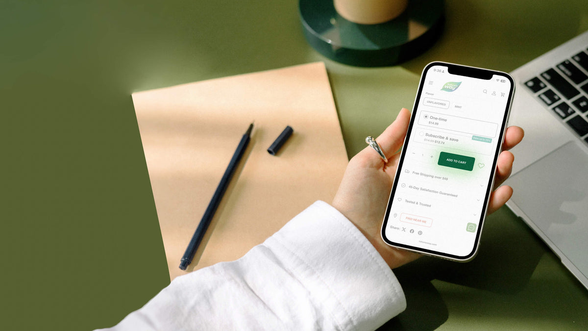A hand holding a smartphone displaying a green and white subscription or payment interface, set against a styled desk background with a notebook, pen, and laptop on an olive green surface.