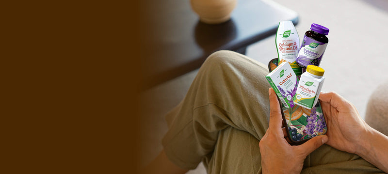 A person sitting indoors holds a bundle of several supplement bottles and boxes in their hands. The products include a white bottle with a purple cap, a white box with green and red graphics, and a purple herbal supplement package.