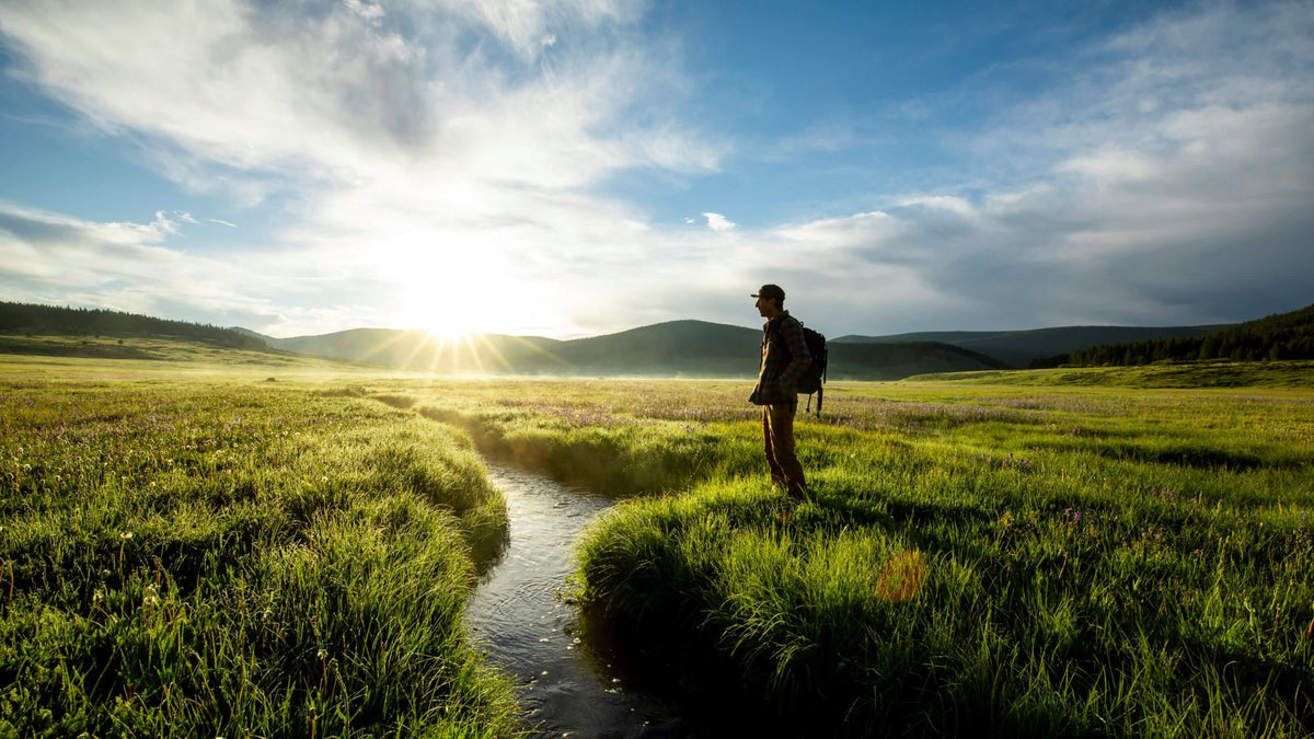 A hiker with a backpack stands on a grassy meadow beside a narrow stream at sunset, with mountains and a bright sun in the sky behind them.