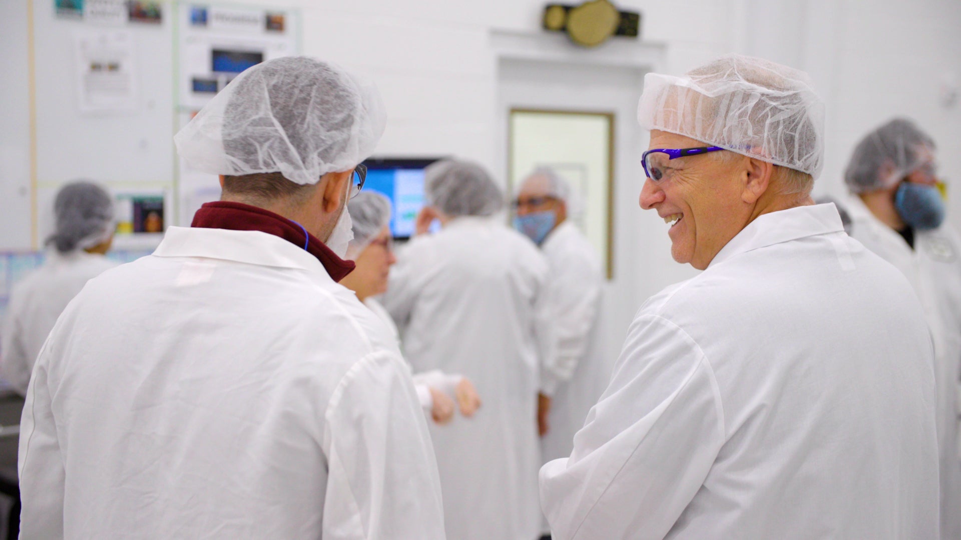 Group of people in a laboratory setting wearing white lab coats and hairnets.
