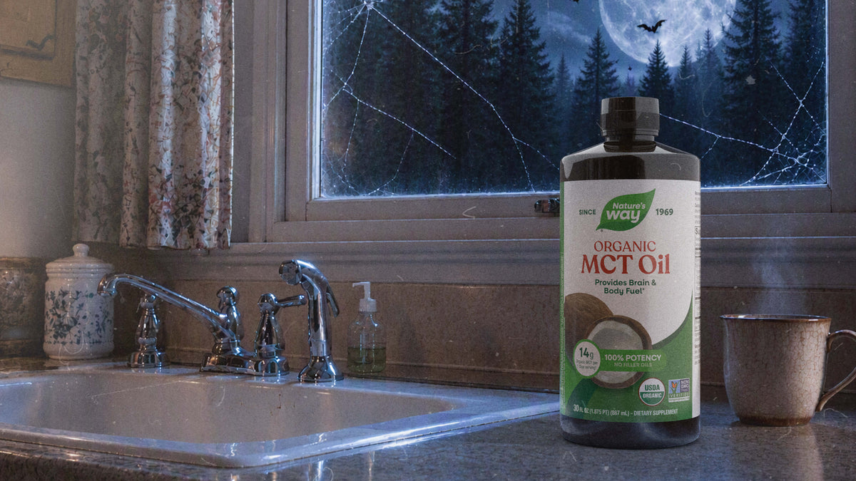 A bottle of Organic MCT Oil stands on a kitchen counter next to a sink, faucet, hand soap dispenser, ceramic jar, and mug, with a window showing a dark forest scene outside.