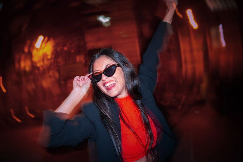 Woman wearing sunglasses and a black jacket with a red top in a dimly lit setting
