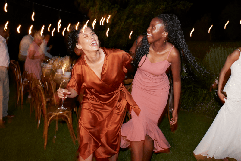 Two women dancing joyfully at an outdoor evening event. The woman on the left wears a shiny rust-orange satin wrap dress and holds a wine glass, while the woman on the right wears a dusty pink sleeveless fit-and-flare dress.