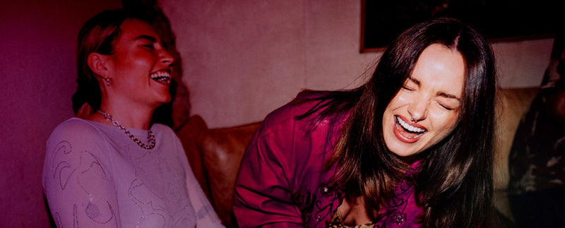 Two women laughing together in a dimly lit room with a pinkish-purple hue.