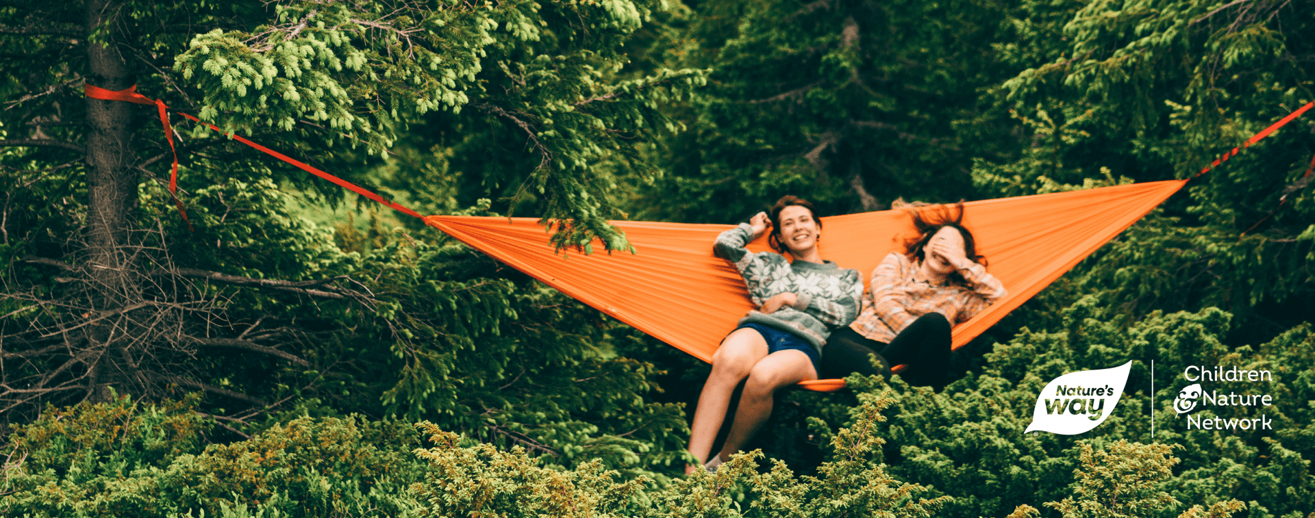 Two women sitting in an orange hammock in the forest