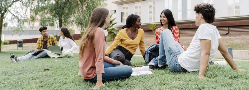 A group of young adults sitting on a grassy lawn talking and laughing outdoors near a building.