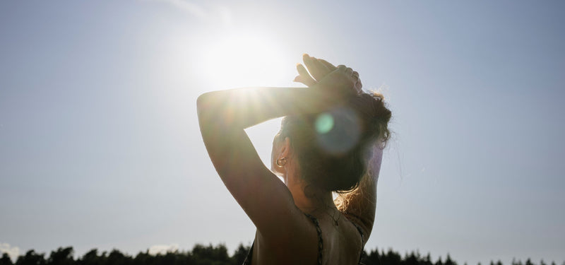 A woman seen from behind with her arms raised, standing outdoors in bright sunlight against a clear blue sky with a tree line in the distance.