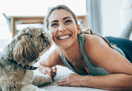 A smiling woman lying with a dog that is playfully sniffing her face