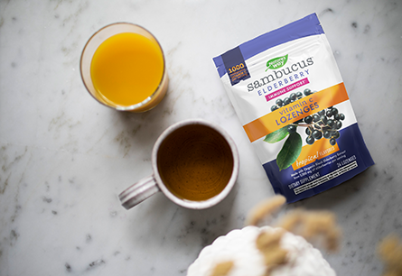 A pouch of Nature's Way Sambucus Vitamin C Lozenges next to a cup of orange juice and mug of tea.