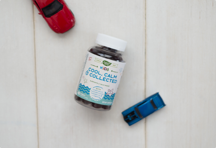 <{%DETAIL1_13369%}>A bottle of Kid's Cool, Calm, & Collected gummies laying between red and blue toy cars on a white table.