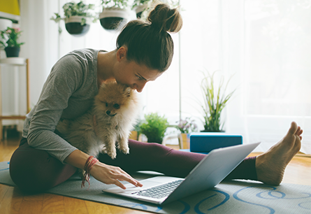 A woman sitting on a yoga mat holding a pomeranian and typing on a laptop with her free hand.