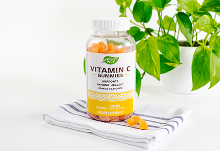 <{%DETAIL1_13607%}>Nature's Way Vitamin C Gummies bottle with 3 gummies outside of the bottle sitting on a kitchen towel on a white surface with Pothos plant in the background 