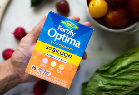 <{%DETAIL1_10337%}>A hand holding a package of Fortify Optima Daily Probiotic next to assorted vegetables.