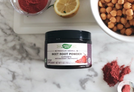 A jar of Beet Root Powder laying on a marble countertop next to small bowls of red powder, a sliced lemon, and a bowl of beans.