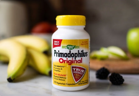 A bottle of Primadophilus Original probiotics sitting on a countertop with berries, bananas, and a sliced green apple in the background.