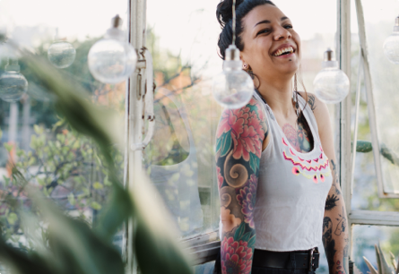 A laughing woman with colorful tattoos on her arms surrounded by plants.